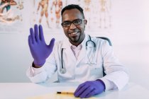 Joven y alegre médico afroamericano en uniforme médico y anteojos sonriendo y agitando la mano hacia la cámara mientras está sentado en la mesa en el laboratorio moderno - foto de stock