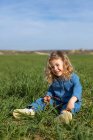 Симпатичный счастливый ребенок, сидящий в зеленом поле в солнечный день, глядя в сторону и играя с травой летом — стоковое фото