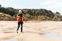 Back view of anonymous aged female backpacker with trekking pole strolling on sandy shore with puddles against green mount — Stock Photo