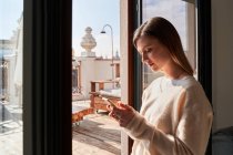 Side view of calm young lady in stylish sweater messaging on smartphone while standing near window at home — Stock Photo