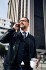 Mature Hispanic male executive in smart casual wear with hot drink to go talking on cellphone while walking on urban pavement — Stock Photo