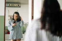 Alegre mujer de mediana edad con blusa a rayas que se refleja en el espejo mientras toma autorretrato en el teléfono inteligente en la habitación en casa - foto de stock