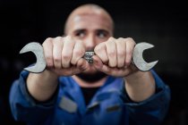 Faceless male mechanic in blue overall covering face with metal spanner in hands looking away on black background — Stock Photo
