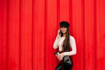 Seriöse stylische Unternehmerin lehnt an roter Wand in der Stadt und diskutiert Projekt auf Smartphone, während sie mit Drink in die Kamera schaut — Stockfoto