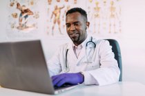 Концентрированный молодой афроамериканский врач-мужчина в медицинском халате и наушниках TWS, работающий на ноутбуке, сидя за столом в современной клинике — стоковое фото