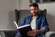 Concentrated young Hispanic guy in casual clothes sitting in comfortable armchair and reading interesting book and looking at camera in daylight at home — Stock Photo