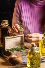 Crop unrecognizable female demonstrating small wooden chest filled with herbs twigs with green leaves near essential oil glass bottles on textile near scissors at table — Stock Photo