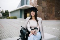 Contenuto imprenditrice in abito alla moda seduta su panchina con tazza di bevanda per andare e distogliere lo sguardo — Foto stock