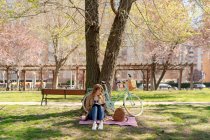 Full body of concentrated young female taking notes in notebook on checkered fabric with backpack near bicycle in park — Stock Photo