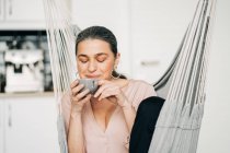 Positive middle aged female with closed eyes drinking hot beverage while sitting on hammock in kitchen with modern kitchenware on blurred background — Stock Photo