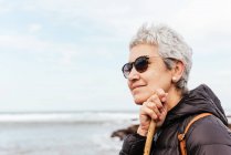 Vue latérale de trekker femme âgée souriante dans des lunettes de soleil avec les cheveux gris regardant loin contre l'océan orageux — Photo de stock