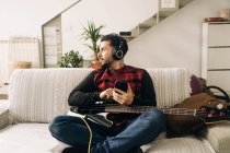 Musicista maschio adulto barbuto con basso chitarra in cuffie messaggistica di testo sul cellulare in soggiorno — Foto stock
