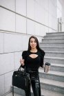 Angolo basso di imprenditrice alla moda con drink da asporto in tazza in piedi vicino a un edificio moderno sulla città e guardando la fotocamera — Foto stock
