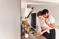 Вид сбоку на молодого романтичного этнического парня в белой футболке, улыбающегося и обнимающего счастливую подругу, готовящую здоровый завтрак с авокадо на кухне — стоковое фото