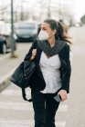 Серйозна жінка в респіраторі з сумкою, що йде по прогулянці зі смартфоном в руці на вулиці біля узбіччя з машинами на розмитому фоні — стокове фото