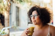 Доросла весела жінка в окулярах, сидячи за міським столом кафетерій зі склянкою напою, дивлячись далеко — стокове фото