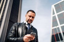 Знизу середнього віку етнічний чоловік-підприємець текстові повідомлення на мобільному телефоні, стоячи в місті — стокове фото