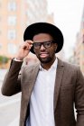 Зміст афроамериканського чоловіка в стильній куртці і капелюсі коригуючи окуляри і дивлячись на камеру стоячи на вулицях міста — стокове фото