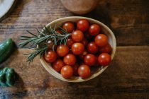 Schüssel mit frischen Kirschtomaten in der Nähe von Rosmarinstielen und ganzen Zwiebeln auf Holztisch — Stockfoto