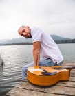 Side view guy in casual clothes sitting with guitar on wooden pier near river with mountains on background under cloudy gray sky in daytime — Stock Photo