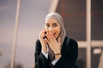 Positivo femminile etnica in hijab in piedi in strada della città e avendo conversazione sul telefono cellulare, mentre guardando altrove — Foto stock