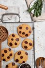 Vista superior de biscoitos doces recém-assados com chips de chocolate na grade de metal colocada na mesa com várias ferramentas de cozinha e ramos de alecrim verde — Fotografia de Stock