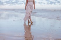 Вид збоку анонімної жінки, що йде у хвилястій воді величезного океану на піщаному пляжі під хмарним небом — стокове фото