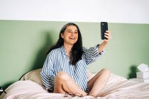 Corpo inteiro de alegre mulher de meia-idade étnica em camisa listrada sorrindo e tirando selfie no smartphone enquanto relaxa na cama durante o fim de semana em casa — Fotografia de Stock