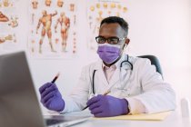 Неузнаваемый серьезный черный ученый в медицинском халате и маске демонстрирует образец крови в пробирке во время видеозвонка на ноутбуке в клинике — стоковое фото
