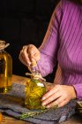 Анонимная женщина в фиолетовом свитере, показывающая эфирные стеклянные бутылки с травами веточки с зелеными листьями рядом с тканью на столе — стоковое фото