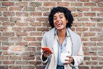 Восхитительная афроамериканка с вьющимися волосами, стоящая с чашкой на кирпичной стене и просматривающая мобильный телефон — стоковое фото