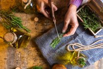 From above of crop lady cutting rosemary sprigs with scissors at table with fabric and rope near essential oil glass bottles and small chest — Stock Photo