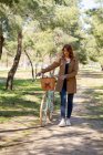 Полное тело молодой женщины, идущей пешком и сконцентрированной возле старого велосипеда с плетеной корзиной в парке — стоковое фото