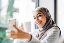 Mulher étnica feliz no hijab e com xícara de bebida tirando selfie no smartphone enquanto desfruta de fim de semana no café — Fotografia de Stock