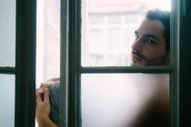 Side view of pensive male embracing knees and sitting on windowsill at home — Stock Photo