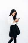 Visão lateral de adolescente etnia feminina em roupa formal em pé perto da parede branca e usando telefone celular — Fotografia de Stock