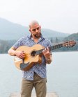 Bearded guy in casual clothes standing with guitar on wooden pier near river with mountains on background under cloudy gray sky in daytime — Stock Photo
