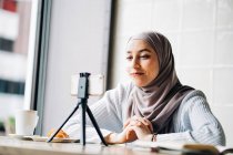 Низкий угол веселой мусульманской женщины в платке съемки видео на смартфоне на штатив для блога, сидя за столом в кафе — стоковое фото