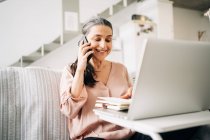 Konzentrierte Frau mittleren Alters telefoniert und macht Notizen im Notizblock, während sie zu Hause auf den Bildschirm des Laptops schaut — Stockfoto