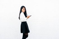 Visão lateral de adolescente etnia feminina em roupa formal em pé perto da parede branca e usando telefone celular e olhando para a câmera — Fotografia de Stock