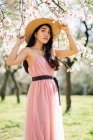 Donna etnica pacifica in cappello di paglia e vestito in piedi sotto i fiori profumati in fiore sui rami degli alberi nel frutteto guardando altrove — Foto stock