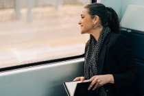 Вид позитивной самки с пончиком, выглядывающим из окна во время чтения книги на пассажирском сиденье во время поездки — стоковое фото