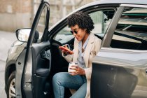 Happy African American female in fashionable sunglasses with thermal mug and using smartphone while smiling and leaving silver prestige automobile — Stock Photo