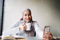 Feliz mujer étnica en hijab y con una taza de bebida tomando selfie en el teléfono inteligente mientras disfruta de fin de semana en la cafetería - foto de stock