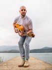 Full body of bearded guy in casual clothes standing with guitar on wooden pier near grass and river with mountains on background under cloudy gray sky in daytime — Stock Photo