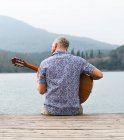 Back view guy in casual clothes sitting with guitar on wooden pier near river with mountains on background under cloudy gray sky in daytime — Stock Photo