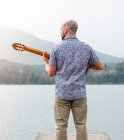 Назад вид бородатий хлопець в повсякденному одязі стоїть з гітарою на дерев'яному пірсі біля річки з горами на фоні під хмарним сірим небом вдень — стокове фото