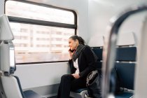 Вид сбоку на женщину, сидящую на пассажирском сиденье и выглядывающую в окно во время телефонного разговора в современном поезде — стоковое фото