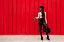 Femme d'affaires confiante dans des vêtements à la mode debout avec des boissons pour aller en ville sur fond de mur rouge et détourner les yeux — Photo de stock