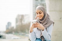 Joven mujer étnica en hijab de pie en la ciudad y mensajería en el teléfono móvil - foto de stock